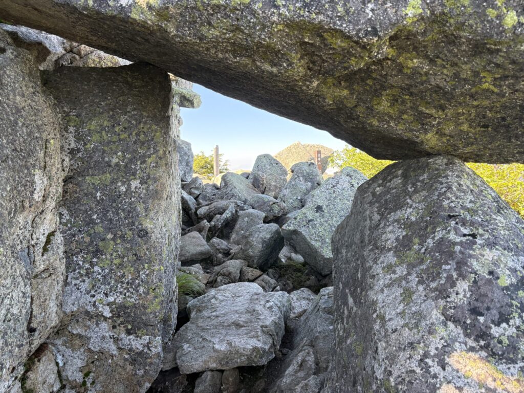 岩のトンネルの向こうに見える山頂標識の画像